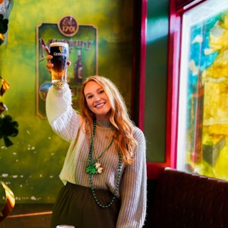 A woman holds up a pint of Guinness in a green bar. She is wearing green and gold necklaces with shamrocks
